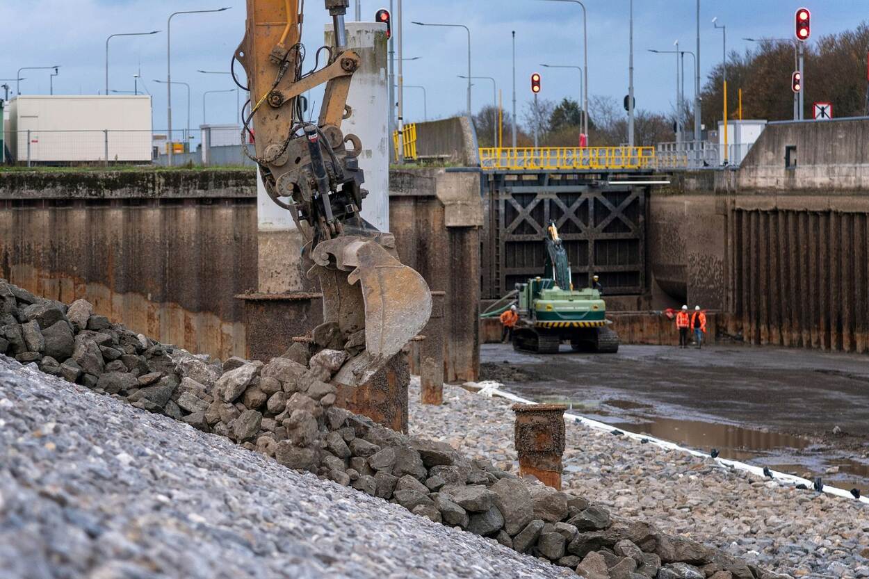 Met graafmachines wordt een steenlaag aangebracht op de betonietmatten. Twee werklui kijken toe.
