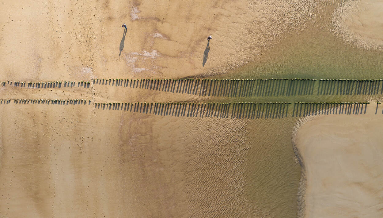 Dronefoto van golfbrekers op een strand dat slechts door twee wandelaars wordt bevolkt