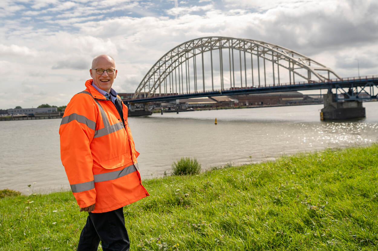 Alwin Nijhuis in een oranje veiligheidsjas staand voor een rivier met een verkeersbrug op de achtergrond