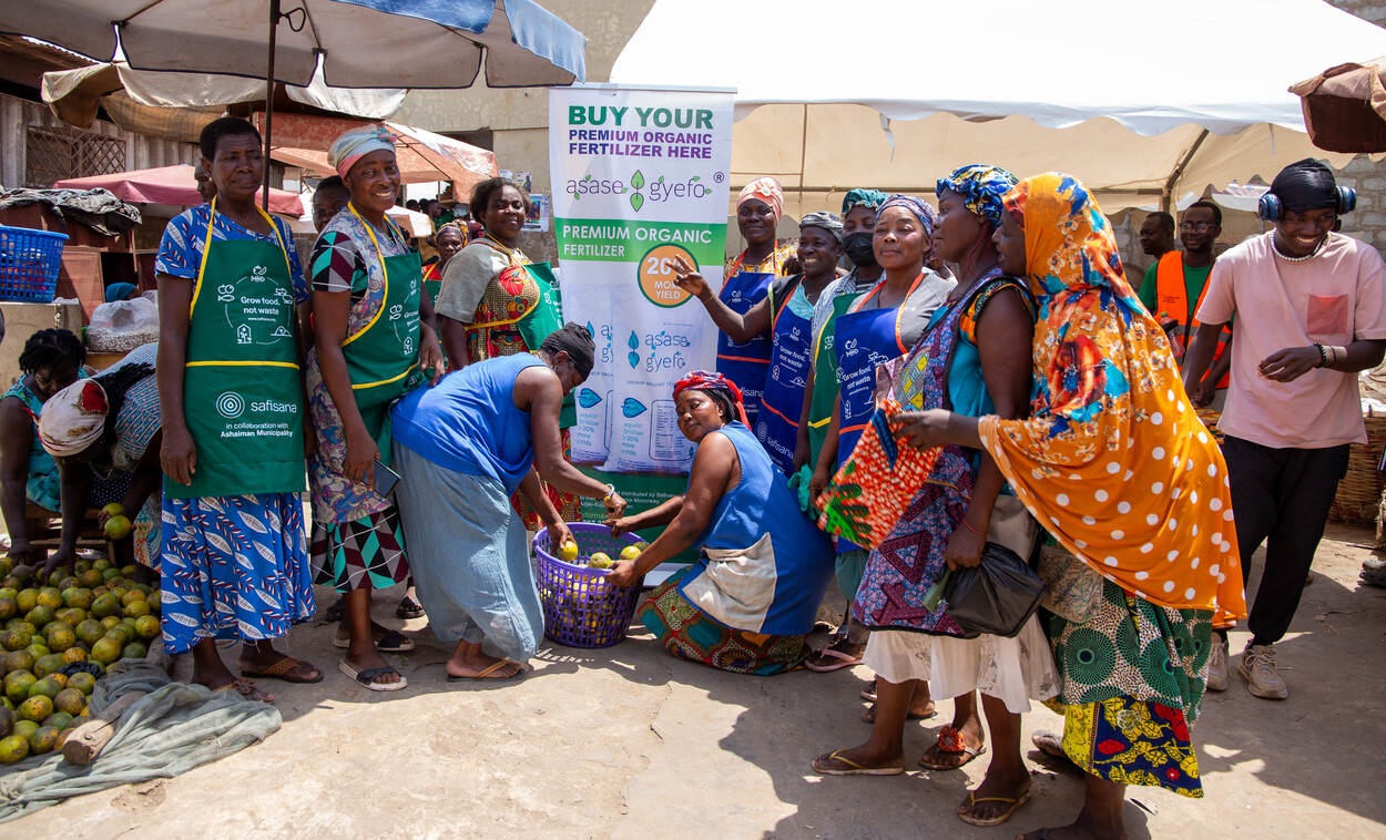 a group of women stand beside a Asaase Gyefo banner at a market