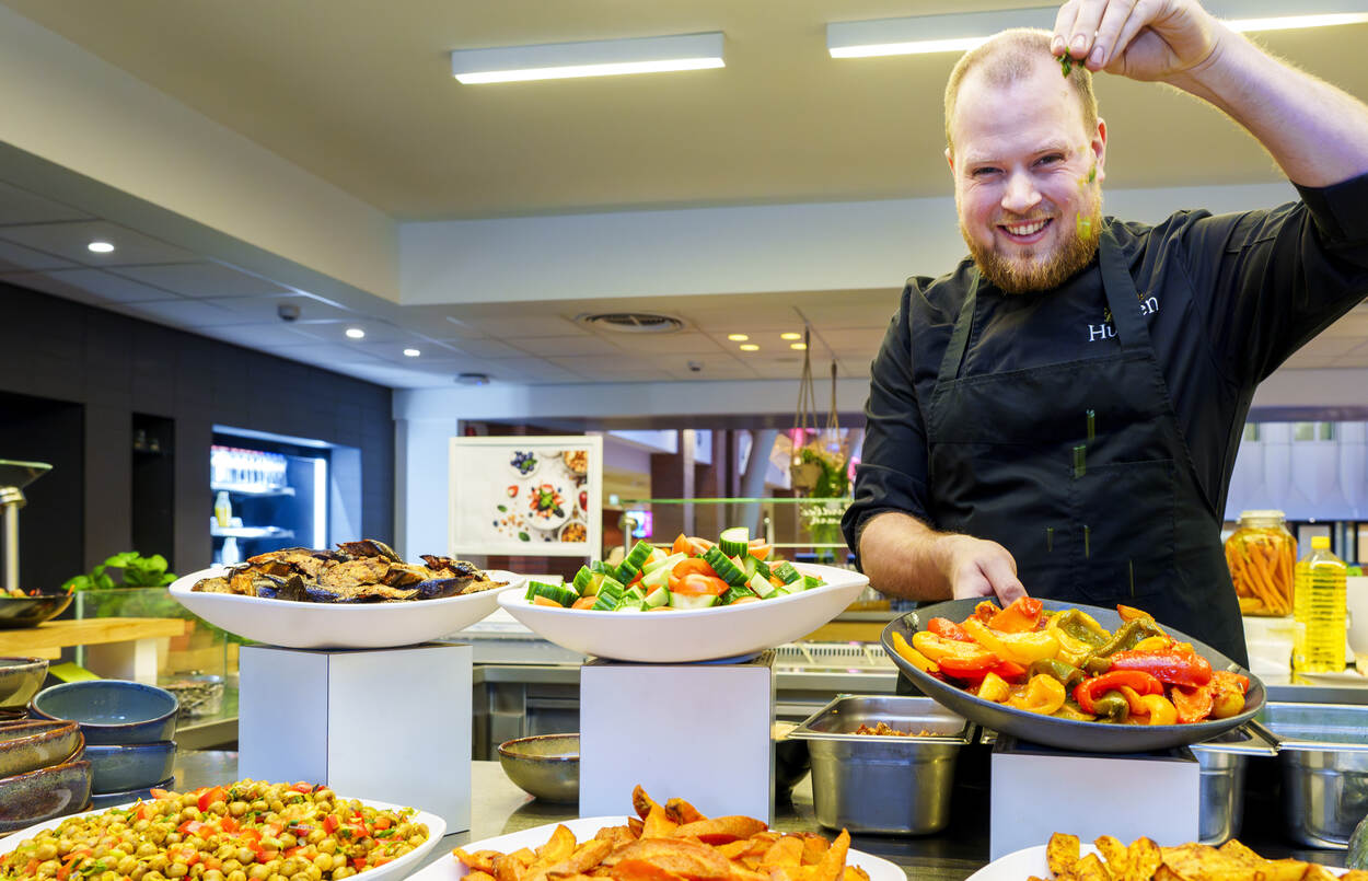 Maarten staat in het bedrijfsrestaurant tussen schalen vol eten en sprenkelt over een schaal nog wat kruiden