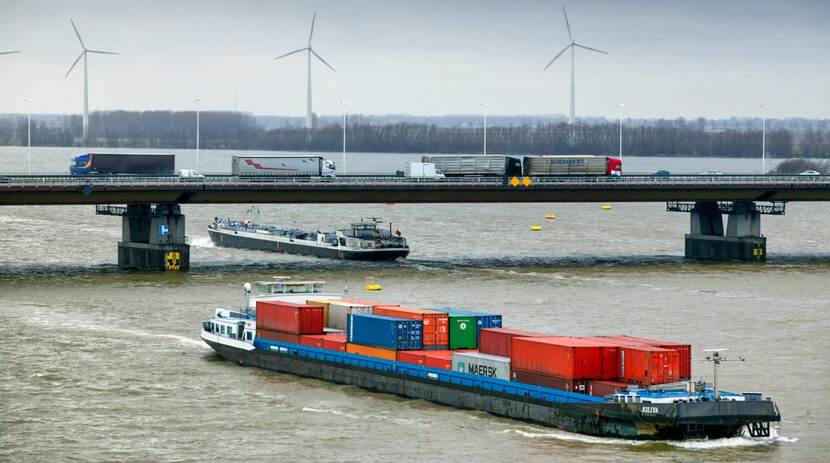 Binnenvaartschip vol containers vaart onder een snelwegbrug met vrachtwagens door.