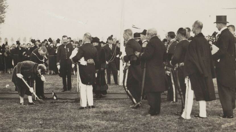 Kroonprinses Juliana bukt zich om met een spade de eerste schop in de grond te zetten, temiddan van een groot gezelschap deftig geklede heren.