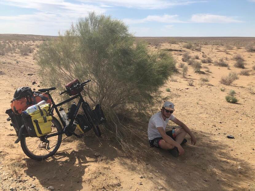 Richard schuilt in de woestijn voor de zon achter een bosje met zijn bepakte fiets naast zich