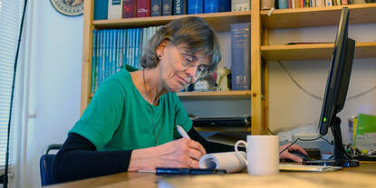 Schrijfster Mieke van Baarsel zit aan tafel en schijft. Op de achtergrond staat een boekenkast