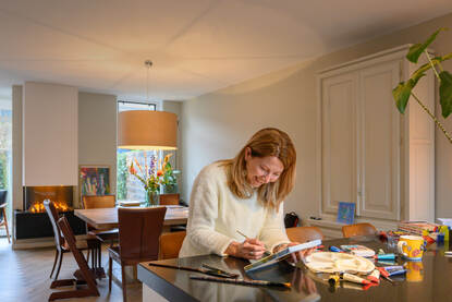Annette Poels zit in haar huiskamer aan tafel te schilderen