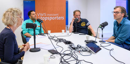 Vier VWS's aan tafel in gesprek voor de opname van een podcast. Op de achtergrond staat een oranje banner met daarop de tekst 'VWS investeert in topsport'