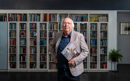 Ad van Liempt voor zijn boekenkast met het boek WO2 in kleur in zijn hand