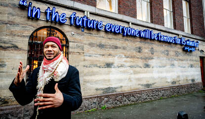 Malcolm staat voor een lichtgevend citaat op een muur: in the future everyone will be famous for 15 minutes.